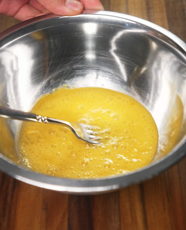 Eggs are fully beaten in a shiny stainless steel bowl using a fork, getting ready to be scrambled for the spam musubi rice bowl.