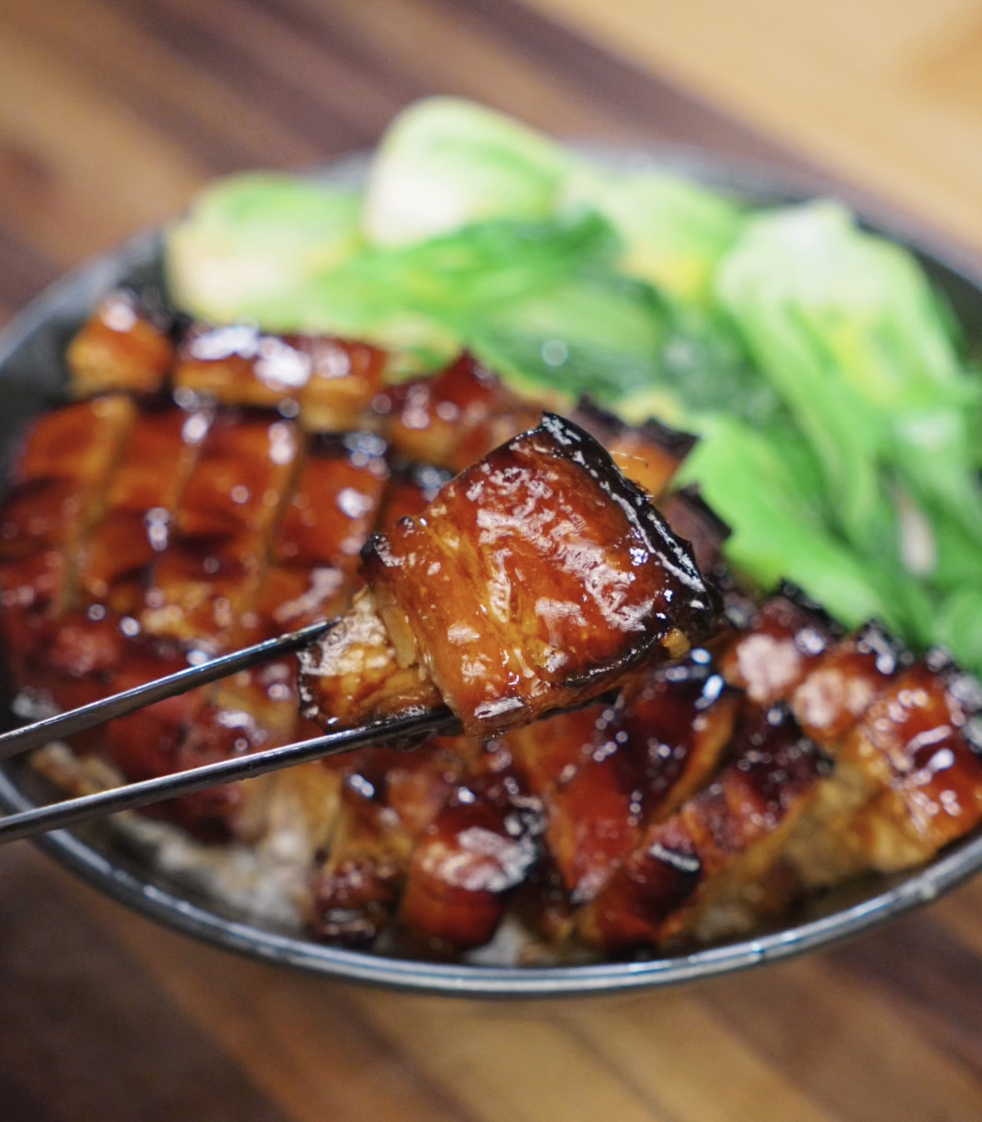 A pair of chopsticks lifts a juicy, glistening bite of honey glazed pork belly from a rice bowl. The caramelized surface is sticky and golden, revealing the tender, juicy layers inside.