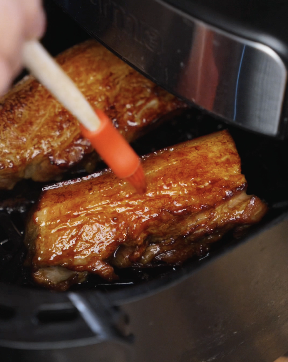 A silicone brush coats sizzling pork belly in the air fryer with more honey glaze. The pork is golden and slightly crisped, ready to soak in more flavor for the final finish.