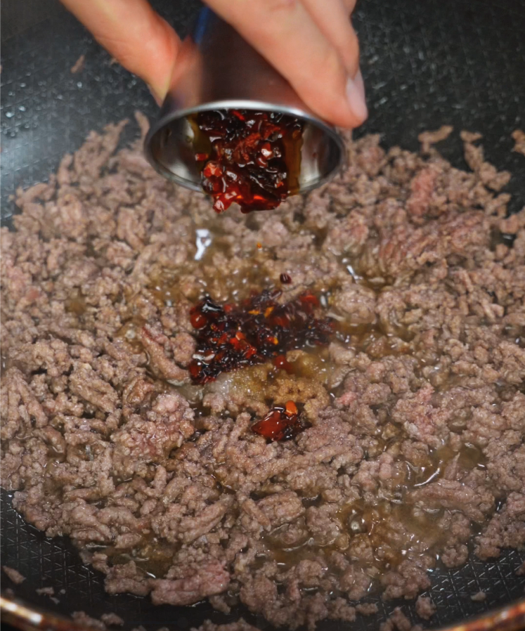 A hand pours spicy chili crisp over sizzling cooked ground beef in a skillet. The chili oil glistens as it hits the meat, infusing it with bold, smoky heat and texture.