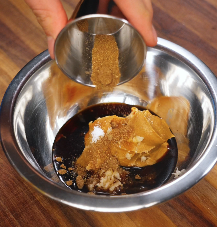 A metal bowl holds a rich blend of seasonings including peanut butter, soy sauce, grated garlic, and brown sugar, while more seasoning is poured in. The mixture is ready to be stirred into a spicy, nutty sauce.