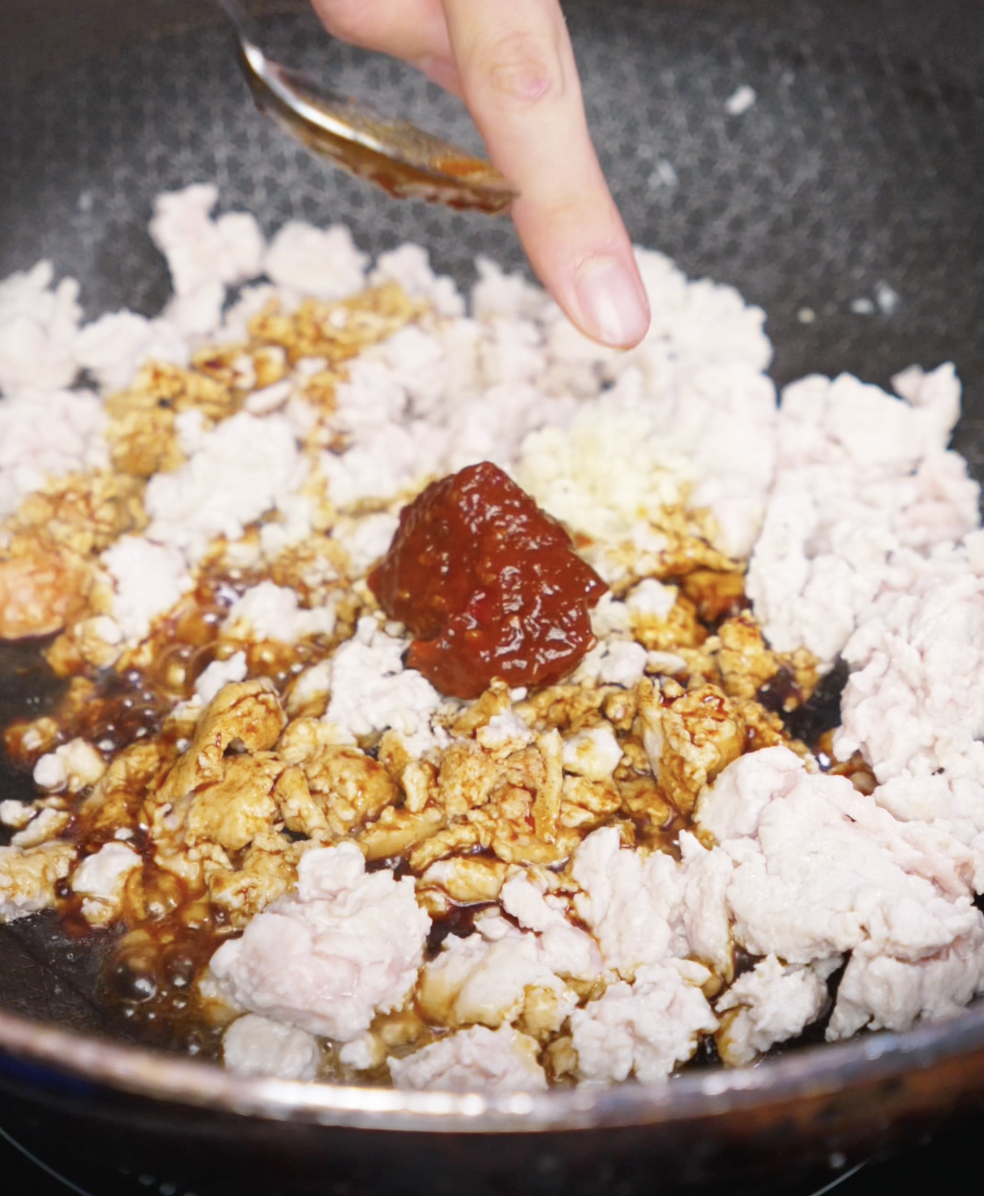 A spoon adds a dollop of soybean paste to partially cooked ground chicken in a pan. The mixture will become the spicy, savory topping for tantanmen noodles.