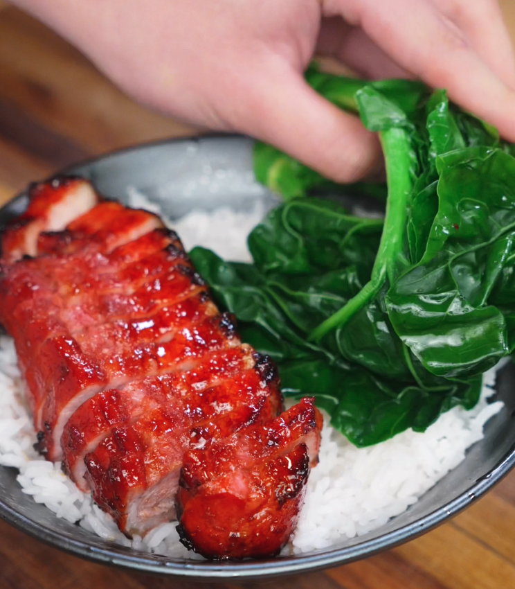 A hand places a bundle of vibrant green vegetables beside char siu pork belly in a rice bowl. The greens balance out the richness of the meat for a complete meal.