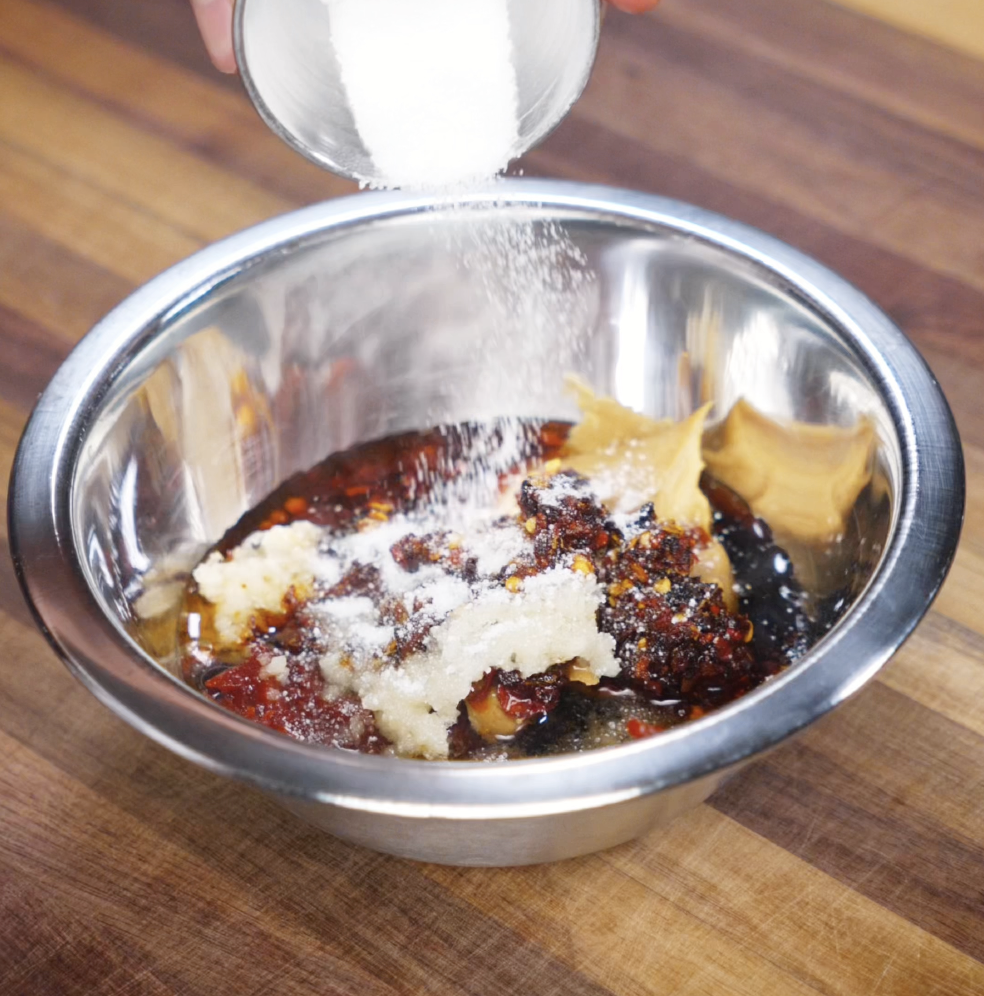 A small bowl of sugar is poured into a mixing bowl filled with gochujang, peanut butter, garlic, and chili oil. The thick, spicy base is being prepped for a bold and creamy sauce.