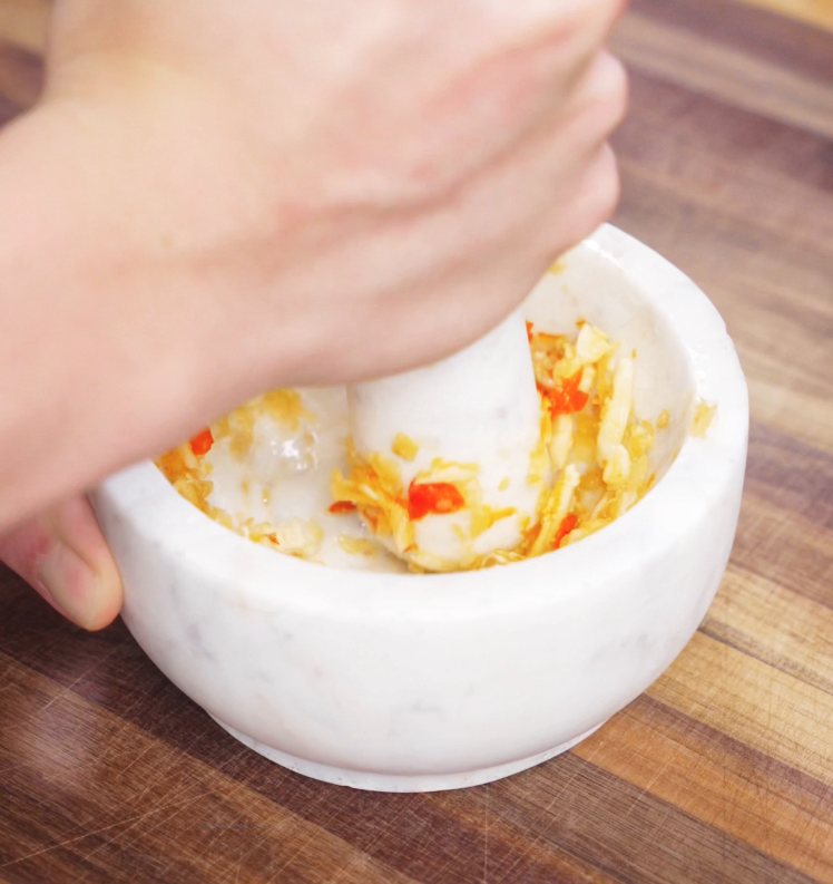 A mortar and pestle filled with crushed garlic and Thai chilis sits on a cutting board. This spicy, aromatic paste adds signature heat and depth to the dish.