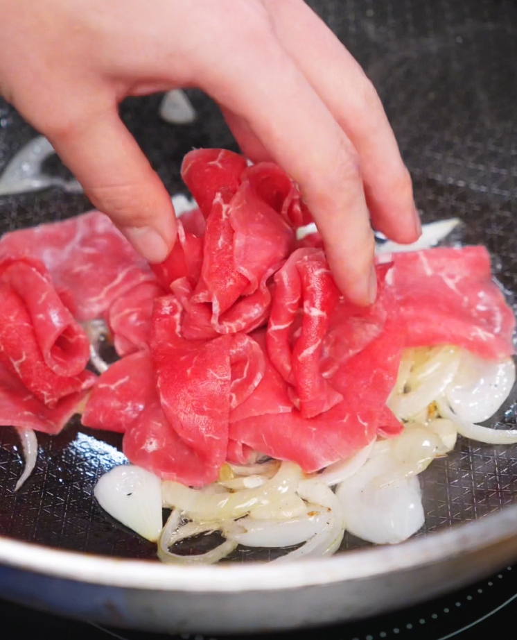 A hand adds a pile of thinly sliced ribeye to sautéed onions in a hot pan. The beef quickly browns while absorbing the savory flavor of the softened aromatics.