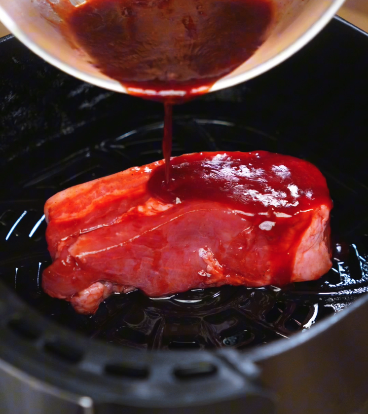 A spoon drizzles reserved char siu marinade over a piece of pork belly inside an air fryer. The glossy sauce caramelizes during cooking, creating a sweet-savory outer crust.