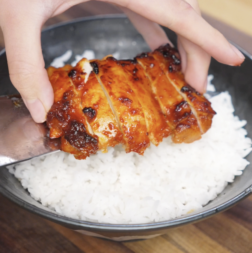 Sliced gochujang-glazed chicken is laid over a bowl of hot white rice. The glossy chicken contrasts with the fluffy rice, forming a hearty and colorful meal base.