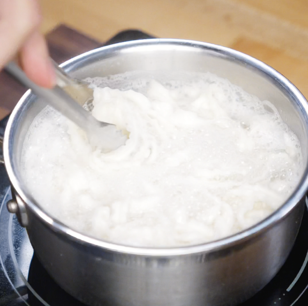A hand uses tongs to stir bubbling instant ramen noodles in a pot of boiling water. The noodles swirl in the starchy water, softening to the perfect chewy texture.