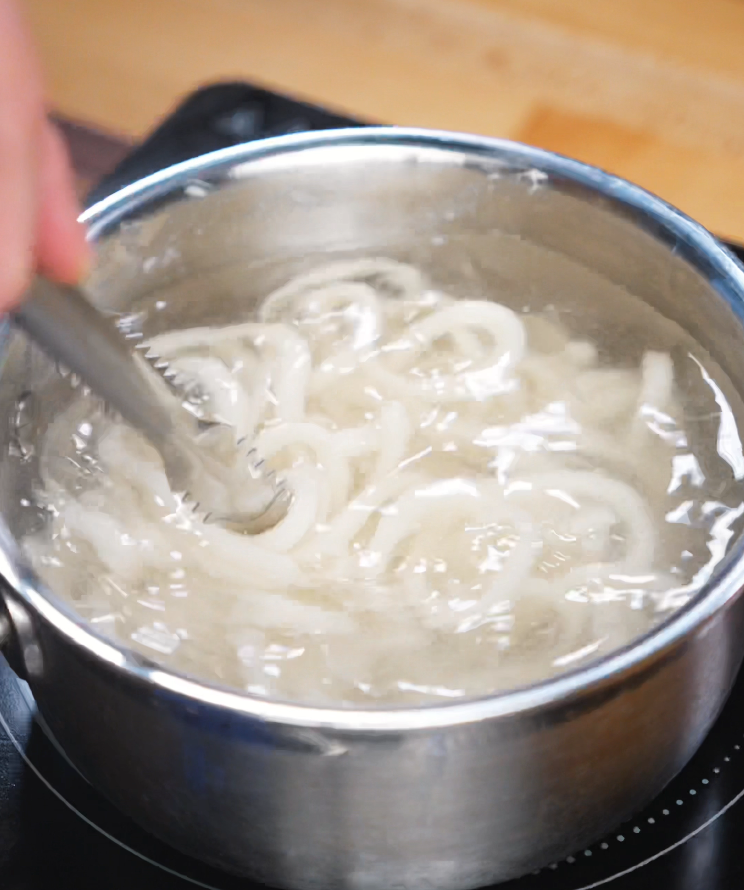 A pot of boiling water bubbles as udon noodles swirl inside. A pair of tongs stirs the noodles to ensure they cook evenly to a chewy, satisfying texture.