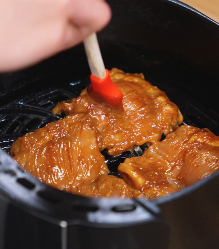 A silicone brush applies more of the rich peanut glaze onto marinated chicken thighs in the air fryer basket, ensuring a deep flavor and beautifully caramelized coating during cooking.