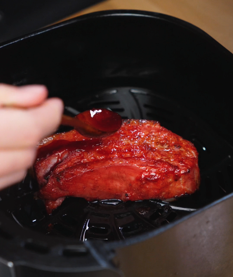 Thick-cut pork belly is coated in bright red glaze and cooks inside an air fryer basket. The edges begin to char slightly, building layers of sticky, smoky flavor.