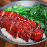 Freshly sliced char siu pork belly rests on top of a bowl of steamed white rice. The glossy glaze glistens under the light, contrasting beautifully with the fluffy grains.