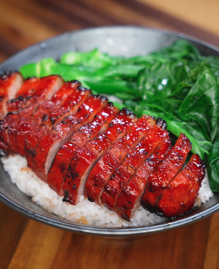 Freshly sliced char siu pork belly rests on top of a bowl of steamed white rice. The glossy glaze glistens under the light, contrasting beautifully with the fluffy grains.