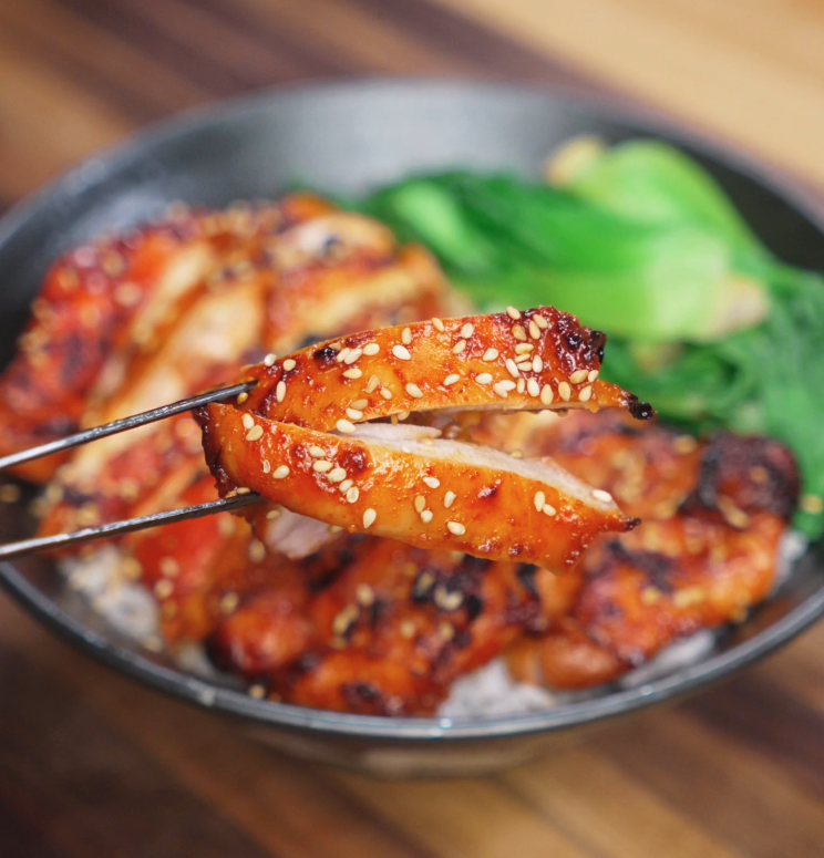 A pair of chopsticks lifts a juicy, glistening bite of gochujang glazed chicken from a rice bowl. The caramelized surface is sticky and golden, revealing the tender, juicy layers inside.