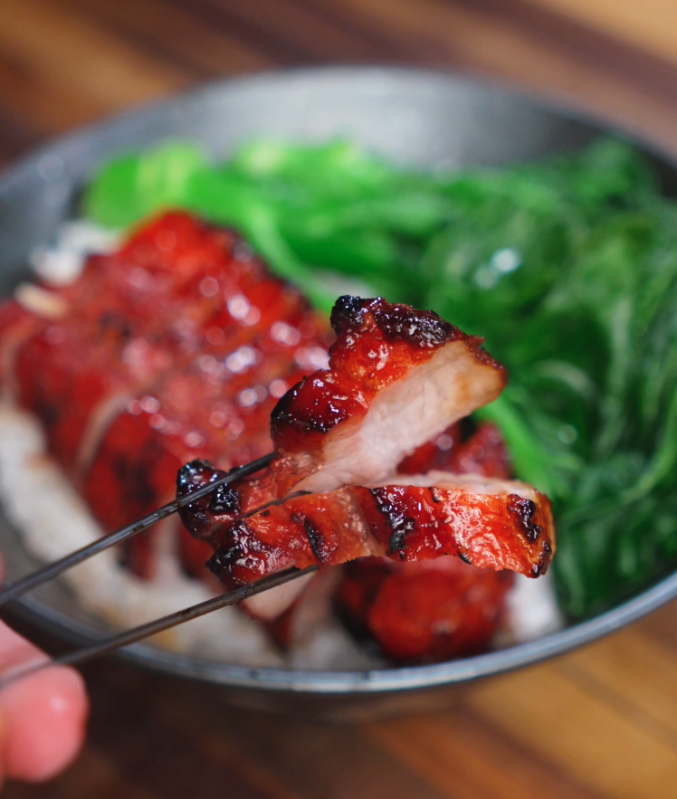 A glossy piece of char siu pork belly is held up with chopsticks. The caramelized edges and juicy center showcase the tender texture and sweet-savory glaze.