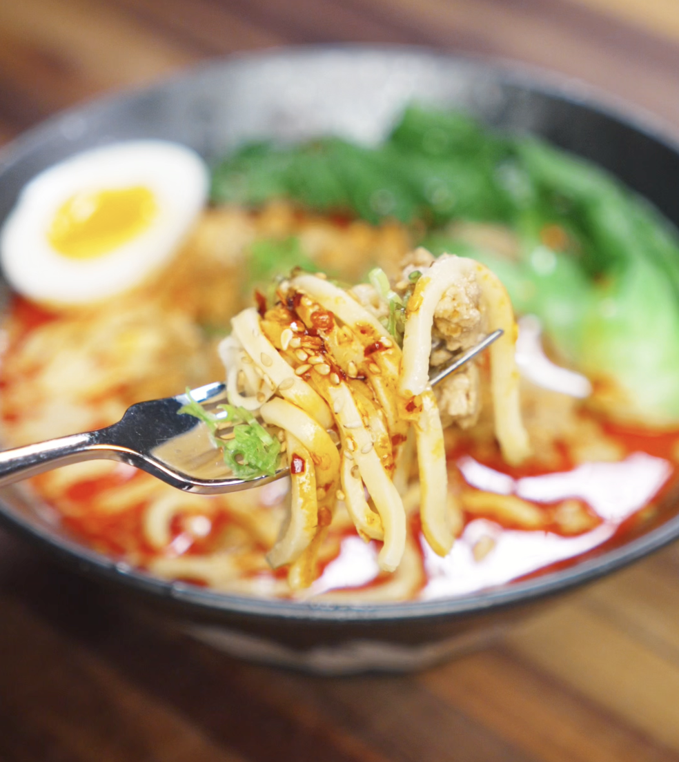 A fork lifts a saucy bite of creamy tantanmen noodles with sesame seeds and scallions. The spicy, rich broth clings to each strand, with tender pork and greens in the background.