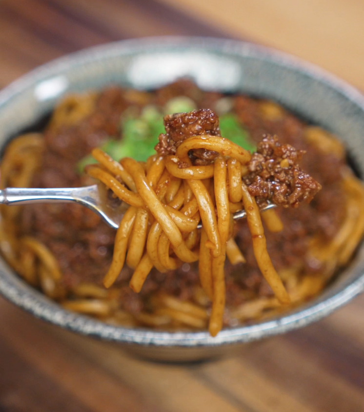 A fork lifts a bite of beefy noodles with crispy garlic bits. The silky noodles and saucy beef showcase layers of umami and garlicky richness.