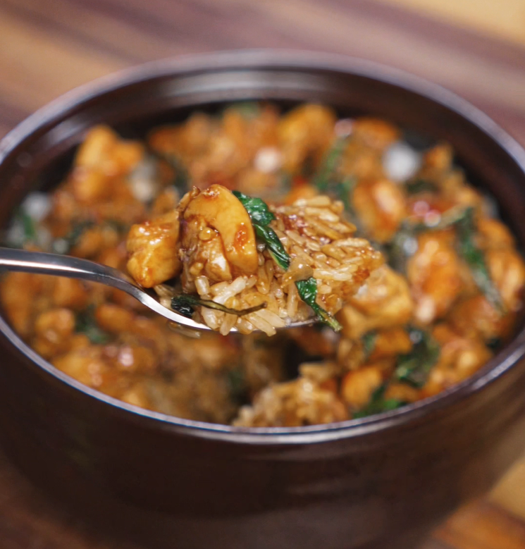 A spoon lifts a bite of Thai basil chicken and rice from the bowl. The glossy sauce, wilted basil, and juicy chicken create a perfect combination of heat, sweetness, and umami.