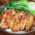 A rice bowl topped with sliced lemon pepper chicken and steamed bok choy. The chicken is golden and caramelized, garnished with sesame seeds for added crunch.