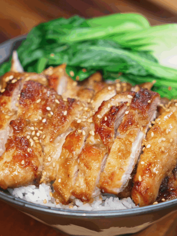 A rice bowl topped with sliced lemon pepper chicken and steamed bok choy. The chicken is golden and caramelized, garnished with sesame seeds for added crunch.