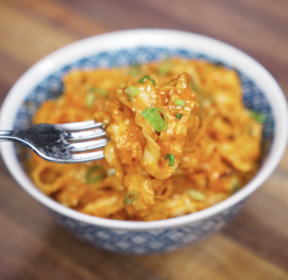 A fork lifts a glossy bite of gochujang peanut noodles from a patterned bowl. The creamy sauce clings to each ribbon-like strand, garnished with scallions and sesame seeds.