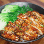 A bowl filled with tender peanut glazed chicken thighs sliced into strips, arranged over a bed of white rice and garnished with scallions and sesame seeds. On the side, bright green steamed bok choy adds freshness and color to the dish.