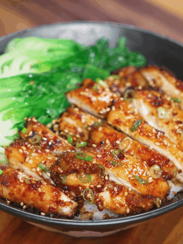 A bowl filled with tender peanut glazed chicken thighs sliced into strips, arranged over a bed of white rice and garnished with scallions and sesame seeds. On the side, bright green steamed bok choy adds freshness and color to the dish.