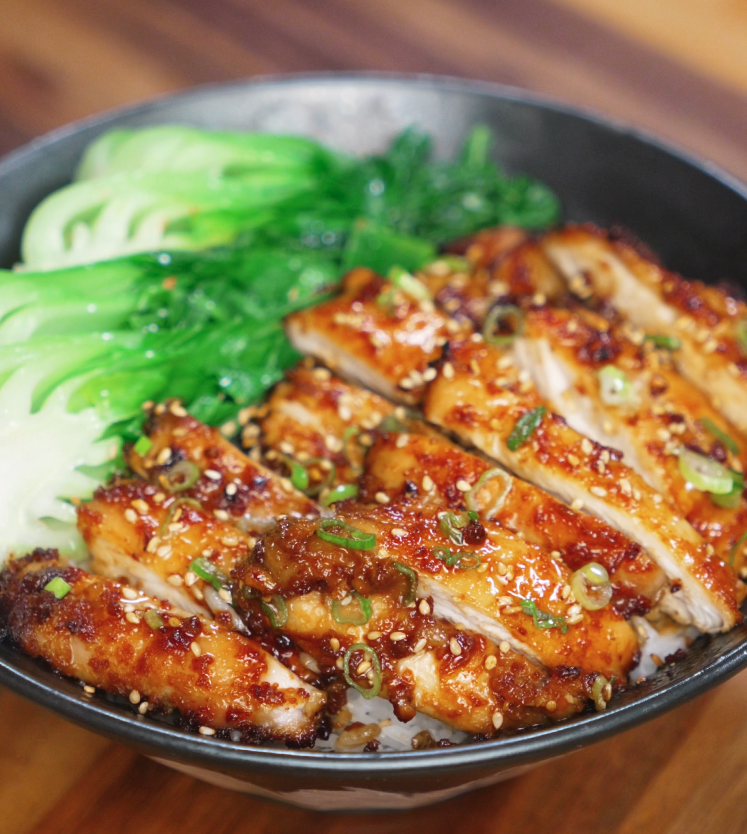 A bowl filled with tender peanut glazed chicken thighs sliced into strips, arranged over a bed of white rice and garnished with scallions and sesame seeds. On the side, bright green steamed bok choy adds freshness and color to the dish.