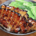 A bowl of white rice topped with glistening, thick-cut slices of honey glazed pork belly. Each piece is lacquered with a caramelized sauce and served alongside vibrant steamed bok choy.