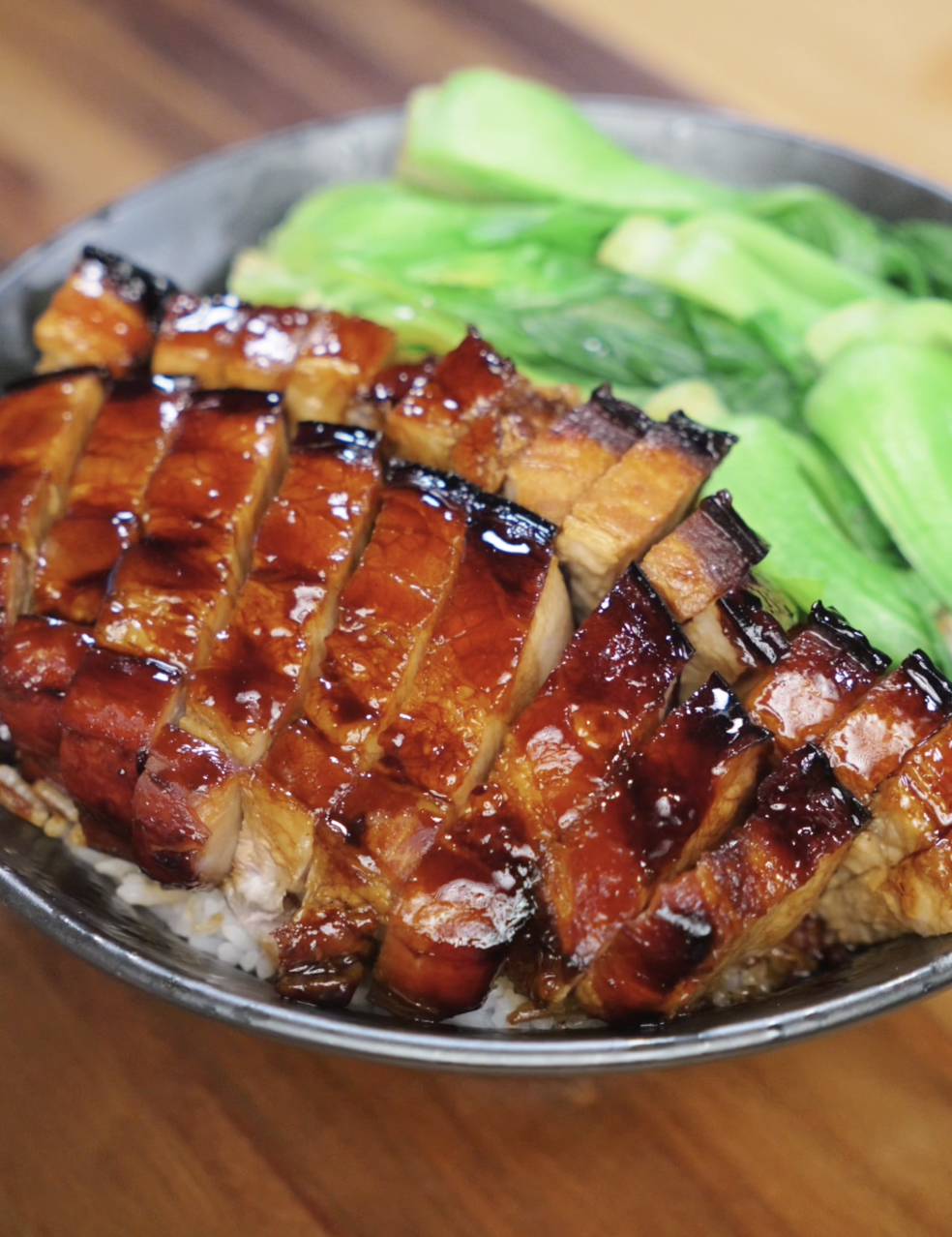 A bowl of white rice topped with glistening, thick-cut slices of honey glazed pork belly. Each piece is lacquered with a caramelized sauce and served alongside vibrant steamed bok choy.