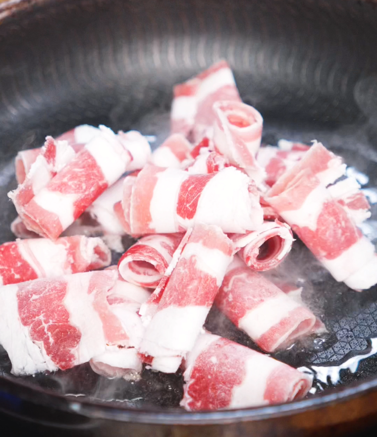A pan filled with tightly rolled slices of frozen beef sizzling as they begin to brown. The marbled ribbons are slowly unfolding and releasing their fat into the hot pan.