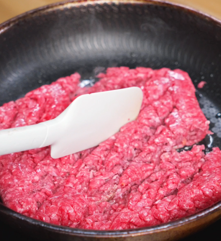 A spatula presses down on a layer of raw ground beef in a hot skillet, beginning the browning process. The beef is vibrant red and sizzling, signaling the start of its transformation into a flavorful base for the BBQ dish.
