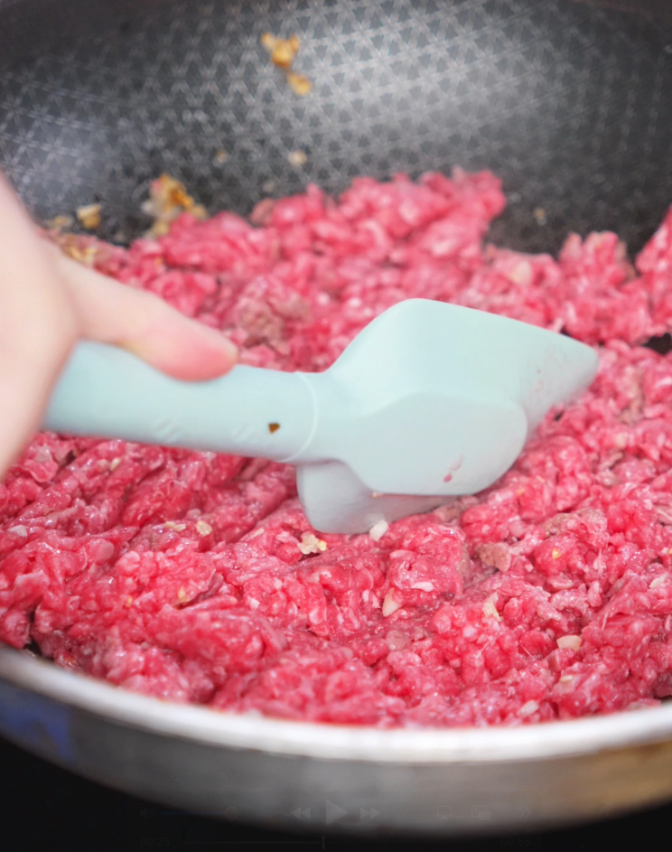 Ground beef is broken apart in a pan with sautéed garlic. The meat starts to brown and soak in garlicky richness.