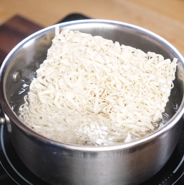 A block of uncooked instant ramen noodles boils in a stainless steel pot filled with hot water, starting to soften and separate.