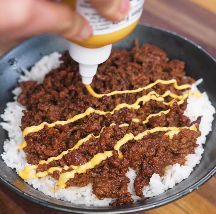 A hand drizzles sriracha mayo in a zigzag pattern over a bed of BBQ beef and white rice. The creamy sauce adds a spicy, tangy finish to the savory dish.