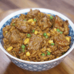 A full bowl of black pepper beef fried rice showcasing glossy chunks of seared beef, golden corn, green peas, and fluffy rice coated in a peppery soy-based sauce. The dish is rich, savory, and meal-prep ready.