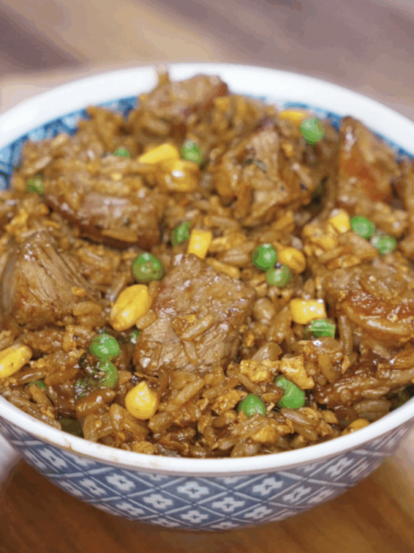 A full bowl of black pepper beef fried rice showcasing glossy chunks of seared beef, golden corn, green peas, and fluffy rice coated in a peppery soy-based sauce. The dish is rich, savory, and meal-prep ready.