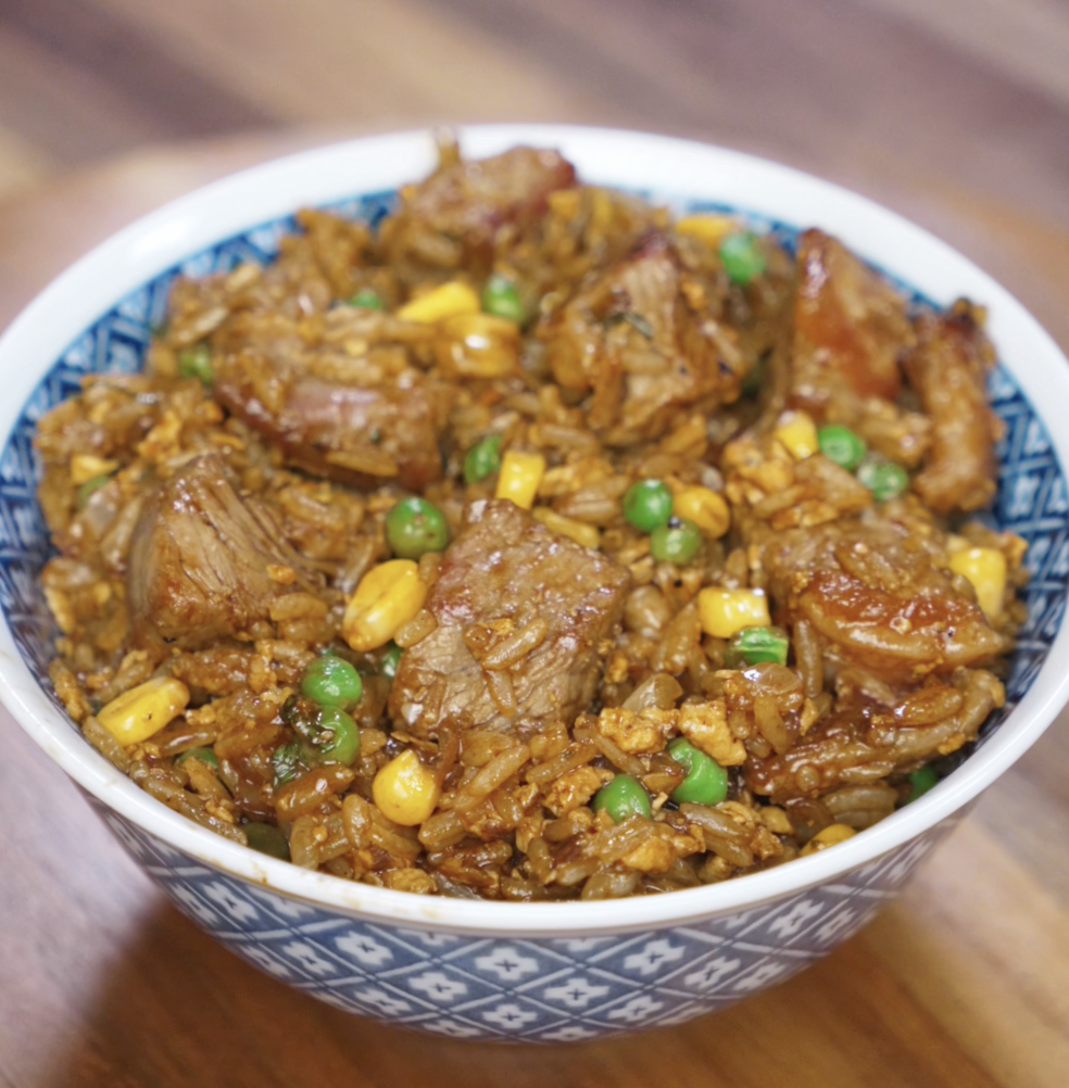 A full bowl of black pepper beef fried rice showcasing glossy chunks of seared beef, golden corn, green peas, and fluffy rice coated in a peppery soy-based sauce. The dish is rich, savory, and meal-prep ready.