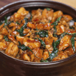 A bowl of Thai basil chicken sits atop a bed of steamed white rice, glistening with savory sauce and flecked with wilted basil leaves. The chicken is glossy, tender, and packed with flavor from garlic, chili, and soy.