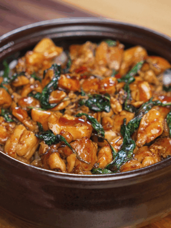 A bowl of Thai basil chicken sits atop a bed of steamed white rice, glistening with savory sauce and flecked with wilted basil leaves. The chicken is glossy, tender, and packed with flavor from garlic, chili, and soy.