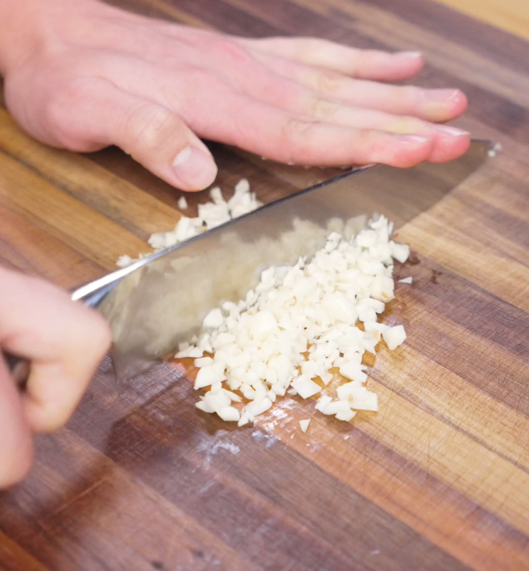 A hand minces garlic finely on a wooden cutting board. The garlic will be used in two stages to infuse both the beef and noodles with bold, aromatic flavor.
