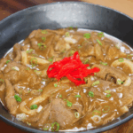 A finished bowl of beef curry udon topped with vibrant pickled ginger and sliced scallions. The thick curry sauce envelops the noodles and beef, ready to be mixed and enjoyed.