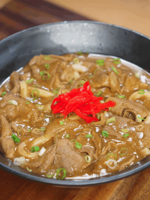 A finished bowl of beef curry udon topped with vibrant pickled ginger and sliced scallions. The thick curry sauce envelops the noodles and beef, ready to be mixed and enjoyed.