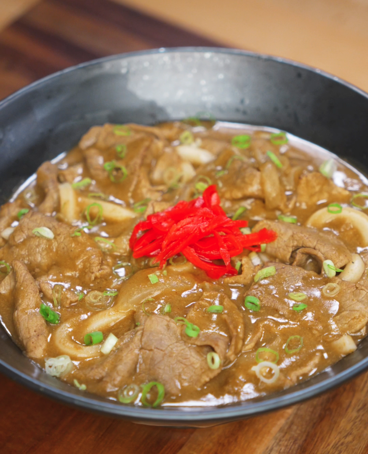 A finished bowl of beef curry udon topped with vibrant pickled ginger and sliced scallions. The thick curry sauce envelops the noodles and beef, ready to be mixed and enjoyed.