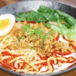 A bowl of creamy tantanmen noodles sits topped with ground meat, sesame chili oil, scallions, bok choy, and a jammy egg. The orange broth is rich, spicy, and velvety.