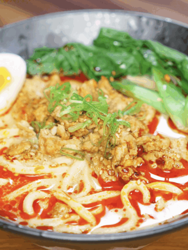 A bowl of creamy tantanmen noodles sits topped with ground meat, sesame chili oil, scallions, bok choy, and a jammy egg. The orange broth is rich, spicy, and velvety.