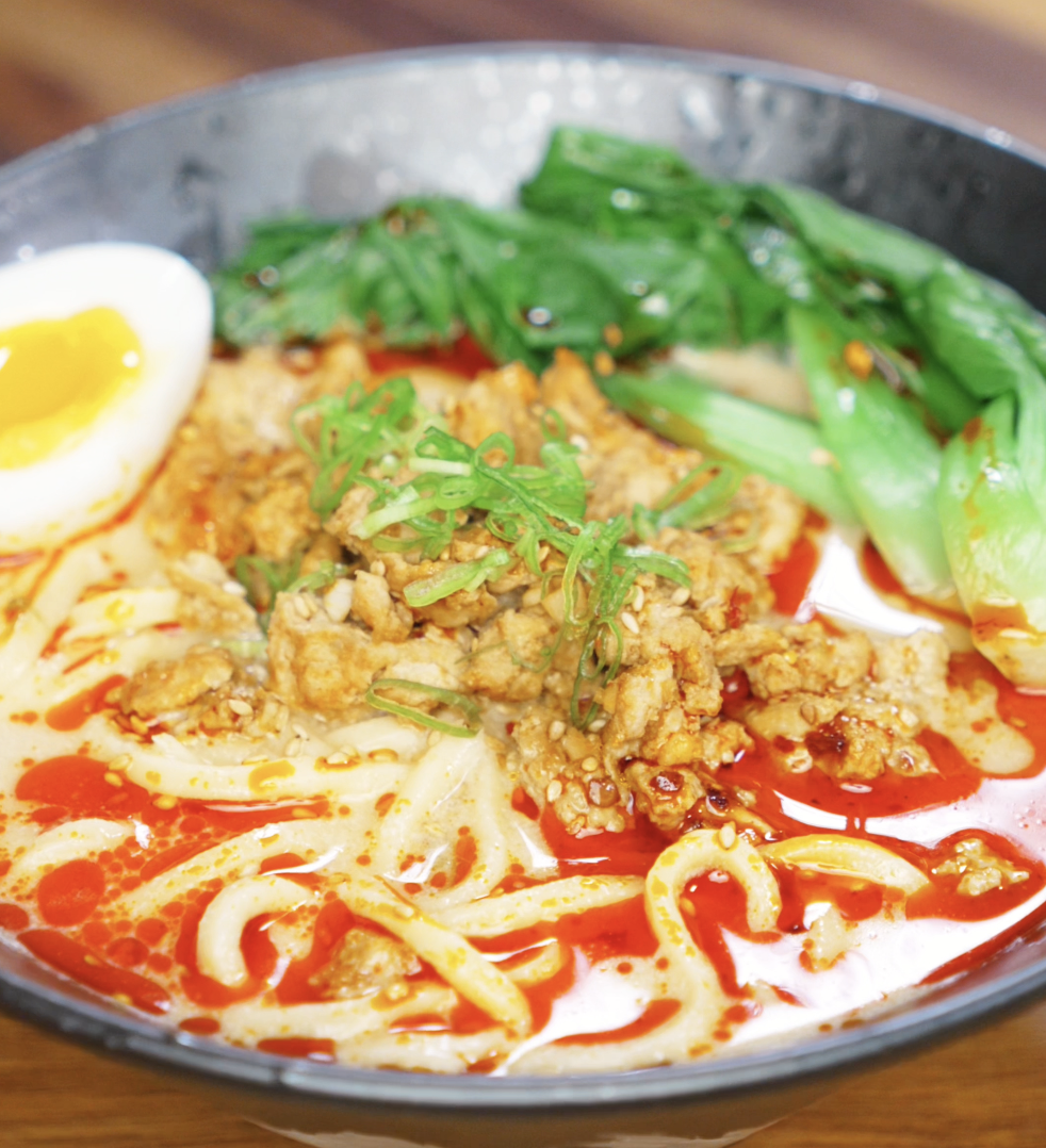 A bowl of creamy tantanmen noodles sits topped with ground meat, sesame chili oil, scallions, bok choy, and a jammy egg. The orange broth is rich, spicy, and velvety.