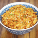 A bowl brimming with glossy gochujang peanut noodles, topped with scallions and sesame seeds. The sauce is rich and orange-red, coating every curl of noodle with bold, nutty flavor.