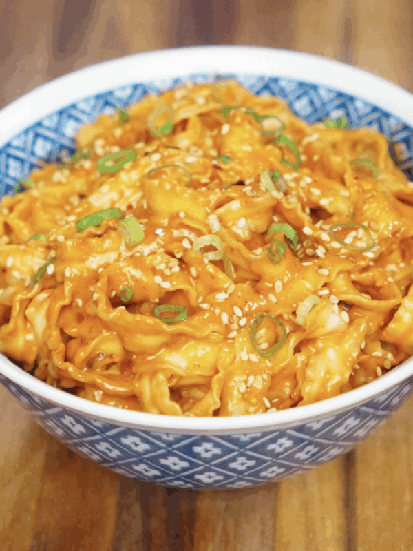 A bowl brimming with glossy gochujang peanut noodles, topped with scallions and sesame seeds. The sauce is rich and orange-red, coating every curl of noodle with bold, nutty flavor.